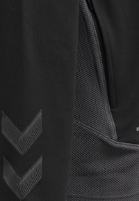 Close-up van zwart textuurstof met twee reflecterende chevron-pijlen en een ritssluitingzak op een kledingstuk.