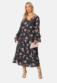 Vestido largo floral con fondo oscuro, mangas acampanadas, dobladillo en capas y escote en V. Combinado con un bolso de mano claro y tacones.
