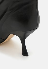 Černá kožená vysoká bota na podpatku s špičatou špičkou, elegantním designem a tenkým, černým strukturovaným stiletto podpatkem.