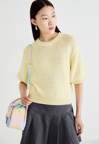 Pull amarelo tricotado com mangas curtas e uma bainha canelada, combinado com uma saia cinza plissada. O modelo segura uma bolsa pastel de várias cores.