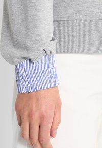 Szary sweatshirt z prążkowanym dołem, z niebiesko-białym paskowanym mankietem wystającym z dołu, podkreślonym marszczonym wzorem.