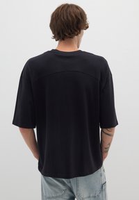 T-shirt oversize in cotone nero con maniche corte e collo rotondo, dotato di una cucitura sulla parte superiore della schiena. Indossato con jeans azzurri chiari.