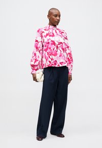 Blusa floral com padrões em rosa e vermelho, gola alta e mangas balão, acompanhada de calças largas em azul-marinho e uma bolsa branca.