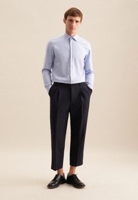 Camicia azzurra chiaro con bottoni e colletto, abbinata a pantaloni scuri a vita alta di colore blu navy. A completare l'outfit ci sono scarpe nere stringate.