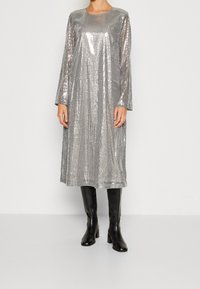 Robe en sequins argentés avec des manches longues, un col rond et une silhouette droite. Portée avec des bottes noires à talons et mi-mollet. Texture lisse.