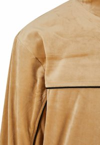 Détail d'une veste en daim camel montrant la couture d'épaule et un passepoil noir le long d'un rabat et de la couture latérale.