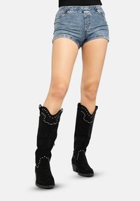 Schwarze kniehohe Stiefel aus Wildleder mit einer gezackten Oberkante, mit Nieten verziert und einem Blockabsatz, kombiniert mit blauen Jeansshorts.