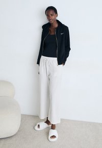 Vrouw staat in een casual outfit met een zwarte rits hoodie, zwart tanktop, witte drawstring broek en witte fluffy slippers tegen een witte muur.