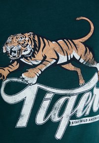 Camiseta de algodón verde con un gráfico impreso de un tigre feroz en naranja y negro, con texto blanco que dice "Tigre" abajo.
