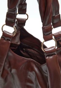 Sac à main en cuir marron avec de larges anses fixées par des anneaux en or, montrant une poche zippée intérieure et un extérieur lisse.