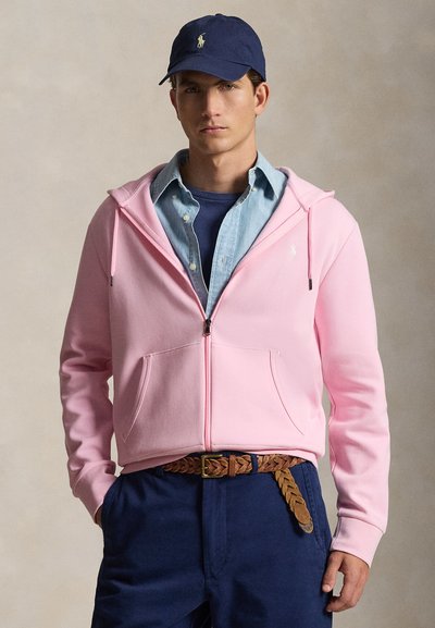 Homme portant une casquette marine, un sweat à capuche zippé rose sur une chemise bleue à boutons, un pantalon marine et une ceinture tressée marron, debout devant un fond neutre.