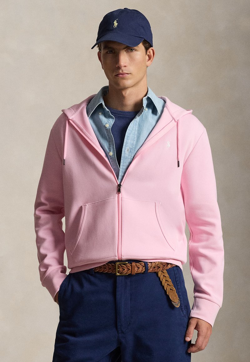 Mees kannab tumesinist mütsi, roosat lukuga kapuutsi sinise nööpide ja kraega särke peal, tumesiniseid pükse ja pruuni kootud vööd, seisab neutraalse tausta ees.