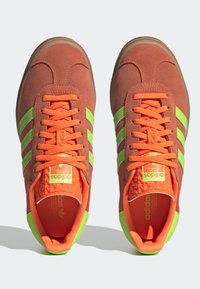 Adidas-sneakers med en överdel i brun mocka, gröna ränder och orange snören. Texturerad inredning och gummisula ger hållbarhet.