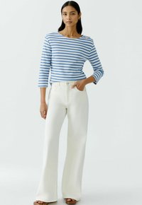 Blau-weißer, gestreifter Langarmshirt, kombiniert mit weißen, weiten Hosen und braunen Plateau-Sandalen. Knopfdetail an der Schulter des Shirts.