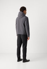 Giacca grigia con cappuccio, realizzata in materiale liscio e resistente all'acqua, abbinata a pantaloni e scarpe neri. Linee pulite, design minimalista.