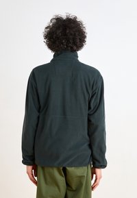 Mörkgrön fleecejacka med hög krage, som har en mjuk textur och en sömdetalj över ryggen. Enkel och funktionell design.
