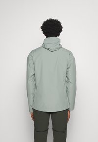 Veste imperméable gris clair avec capuche, dotée d'une texture lisse et de poignets élastiques, portée sur un pantalon vert foncé. Vue arrière affichée.