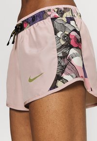 Pantalones cortos deportivos de color rosa claro con un patrón floral en el lado izquierdo, cinturilla elástica y cordón, con un logotipo verde de Nike.