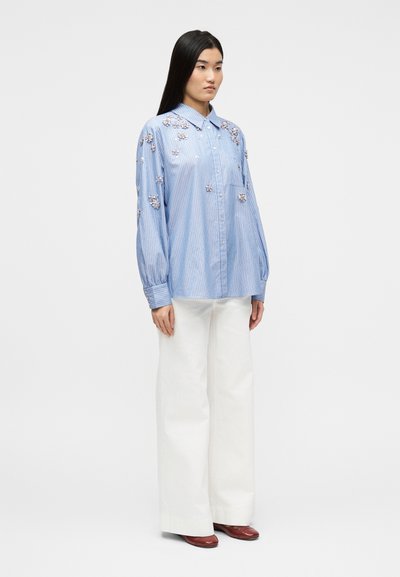 Γυναίκα που στέκεται φορώντας μπλούζα με μπλε ρίγες και ανθοκομικές διακοσμήσεις, φαρδύ λευκό παντελόνι και καφέ παπούτσια, μπροστά σε απλό φωτεινό φόντο.