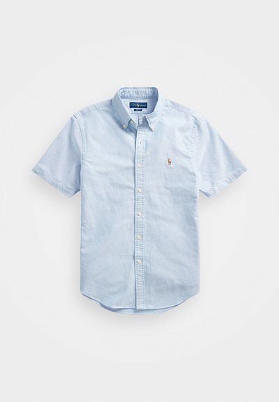 Chemise à manches courtes bleu clair en tissu coton. Elle présente un col boutonné, des boutons sur le devant et un logo brodé sur la poitrine.