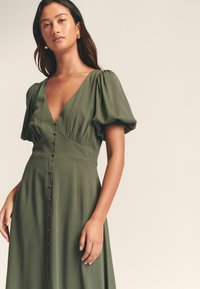 Robe verte olive avec un décolleté en V profond, des manches bouffantes et des détails de boutons sur le devant. Tissu lisse et fluide avec une coupe décontractée.