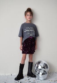 Graues übergroßes T-Shirt mit grafischem Text, kombiniert mit einem glitzernden lila Paillettenrock. Schwarze kniehohe Schnürstiefel. Zwei Discokugeln daneben.