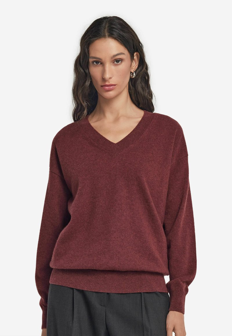 Bordeauxfarbener V-Ausschnitt-Pullover aus weichem, gestricktem Material mit überschnittenen Schultern und lockerer Passform, kombiniert mit maßgeschneiderten, dunklen Hosen.