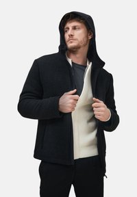 Uomo che indossa una giacca nera con cappuccio sopra un maglione bianco a maglia, tiene la giacca aperta, guardando di lato con un'espressione neutra.