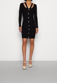 Abito a coste nero con maniche lunghe, scollatura a V profonda e chiusura con bottoni. Presenta spalline sovrapposte. Modella indossa scarpe con piattaforma beige.