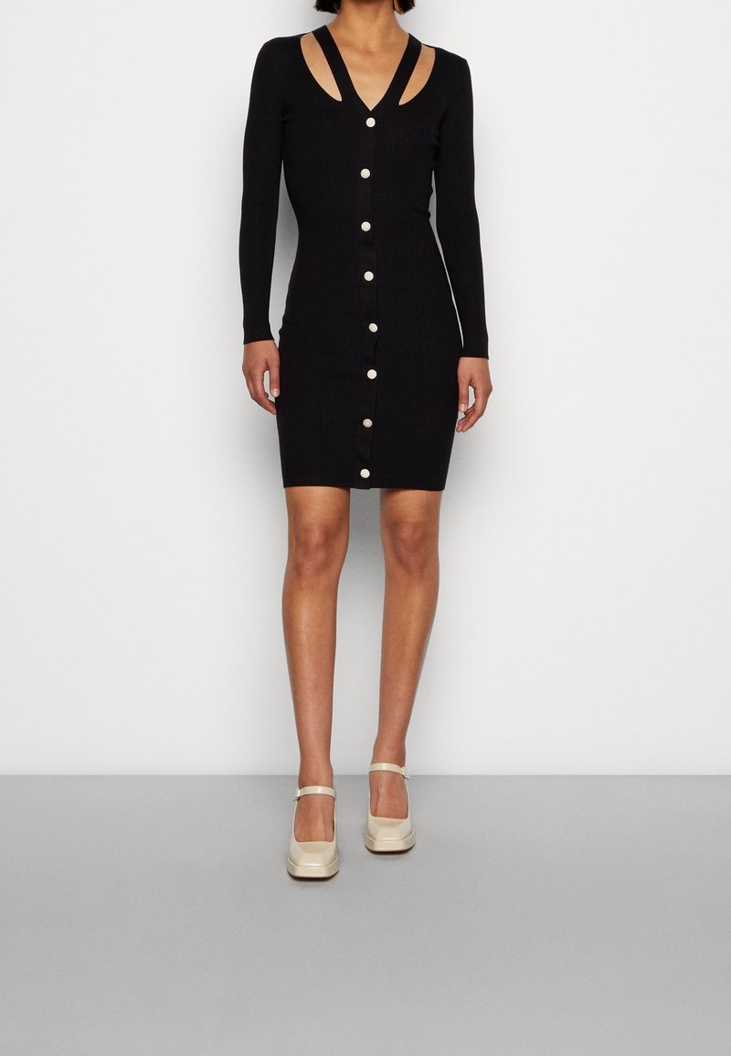 Abito a coste nero con maniche lunghe, scollatura a V profonda e chiusura con bottoni. Presenta spalline sovrapposte. Modella indossa scarpe con piattaforma beige.