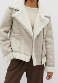 Béžová bunda z umělého beránka s bílým kožíškovým límcem, zapínáním na zip a bočními kapsami. Měkká textura a volný střih. Doplněno hnědým lemováním.