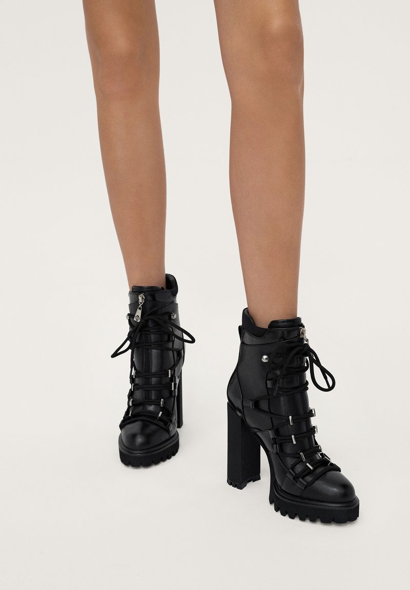 Bottines noires à talons hauts à lacets avec semelles épaisses portées sur des jambes nues sur un fond uni.