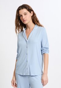 Parte superior de pijama azul claro con mangas largas, botones al frente y ribete blanco a lo largo del cuello y la tapeta, hecha de tela suave y lisa.