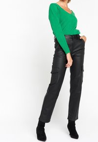 Kvinna klädd i en grön långärmad tröja, svarta högmidjade läderbyxor med fickor och svarta klackade ankelstövlar.