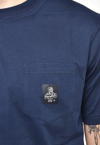 Mörkblå bomullströja med bröstficka och en svart etikett med en hundgrafik och varumärkesnamn, placerad på fickområdet.