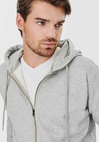 Szara bluza z kapturem na zamek błyskawiczny, z kapturem ze sznurkiem, o miękkiej teksturze i kieszeniach z przodu. Nosi się ją na białej koszulce V-neck, z minimalnym logo.