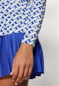 Long-sleeve top with blue geometric pattern and white logo, paired with a pleated blue skirt. Both items feature a smooth texture.