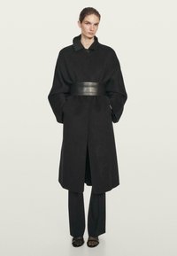 Kvinde iført en lang sort frakke med oversize ærmer, et bredt læderbælte, sorte bukser og sorte sandaler med åbne tæer, stående mod en ensfarvet baggrund.