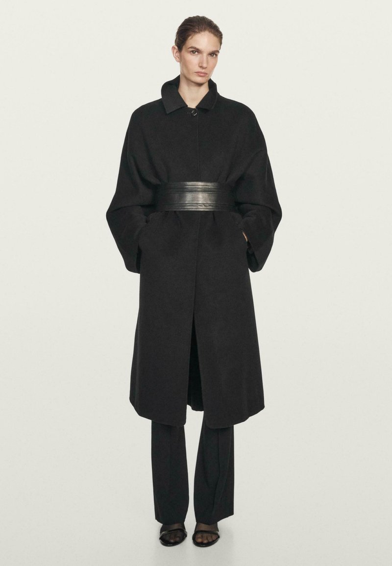 Kvinde iført en lang sort frakke med oversize ærmer, et bredt læderbælte, sorte bukser og sorte sandaler med åbne tæer, stående mod en ensfarvet baggrund.