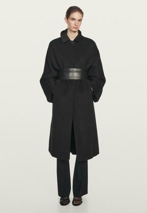 Kvinde iført en lang sort frakke med oversize ærmer, et bredt læderbælte, sorte bukser og sorte sandaler med åbne tæer, stående mod en ensfarvet baggrund.