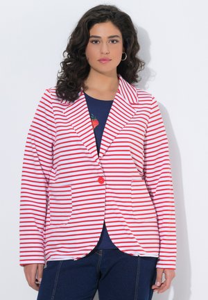 Vrouw met krullend donker haar draagt een rood-wit gestreepte blazer over een marineblauw shirt en een blauwe spijkerbroek, staand tegen een witte achtergrond.