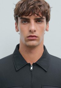 Black zip-up jacket with a collar, featuring a textured material and a chest pocket, worn by a young adult with straight brown hair.