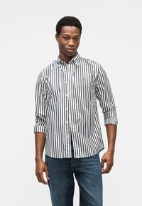 Πουκάμισο με ρίγες σε μαύρο και λευκό, με κλασικό γιακά, μανίκια που γυρίζουν και μικρή λεπτομέρεια λογότυπου στο στήθος. Τζιν παντελόνι.