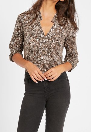 Mujer con blusa de estampado de leopardo con las mangas remangadas metida en unos vaqueros negros de cintura alta, manos entrelazadas en la cintura.