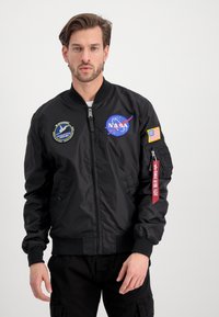 Black bomber jacket with NASA and Smithsonian patches, zippered front, ribbed cuffs, and a fabric tag on the sleeve. Smooth material, casual fit.