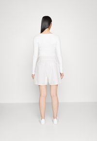 Haut blanc à manches longues associé à un short beige clair, avec une taille élastique. Le mannequin porte des baskets blanches. Design minimaliste.