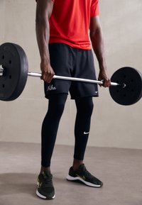 Crne Nike kratke hlače s logom, uske crne tajice, crvena atletska majica i crno/zeleni Nike tenisice. Drži srebrni uteg s crnim težinama.