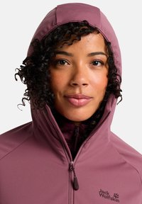 Giacca a cappuccio viola con zip frontale, materiale morbido e logo Jack Wolfskin sulla parte sinistra. Cappuccio aderente.
