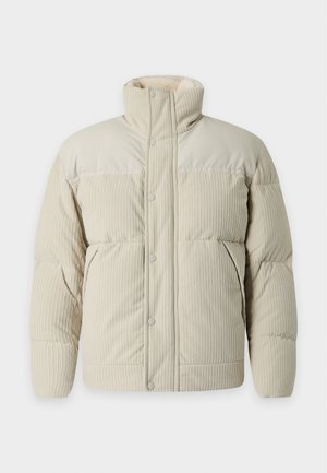 Jeune homme portant une doudoune beige par-dessus une chemise blanche, un jean gris foncé et des baskets noires, debout avec une main dans la poche.