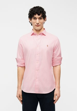 Mann med krøllete mørkt hår iført en lyserosa stripete skjorte med knapper og ermer rullet opp til albuene, står mot en hvit bakgrunn.